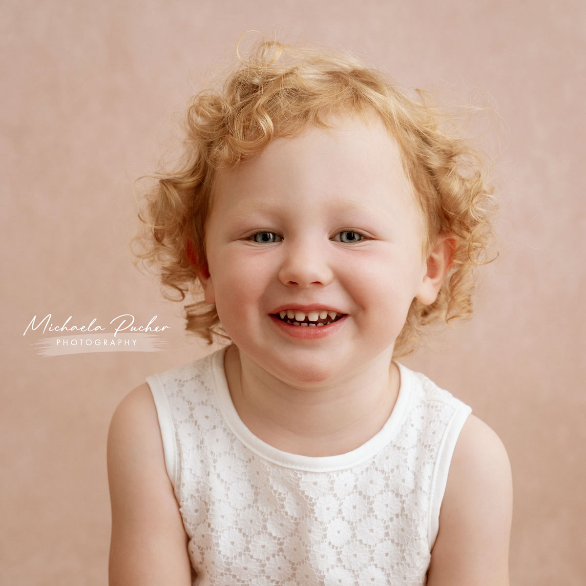 Kleines blondes Mädchen mit lockigen Haaren lacht fröhlich in die Kamera, vor einem zarten roséfarbenen Hintergrund.